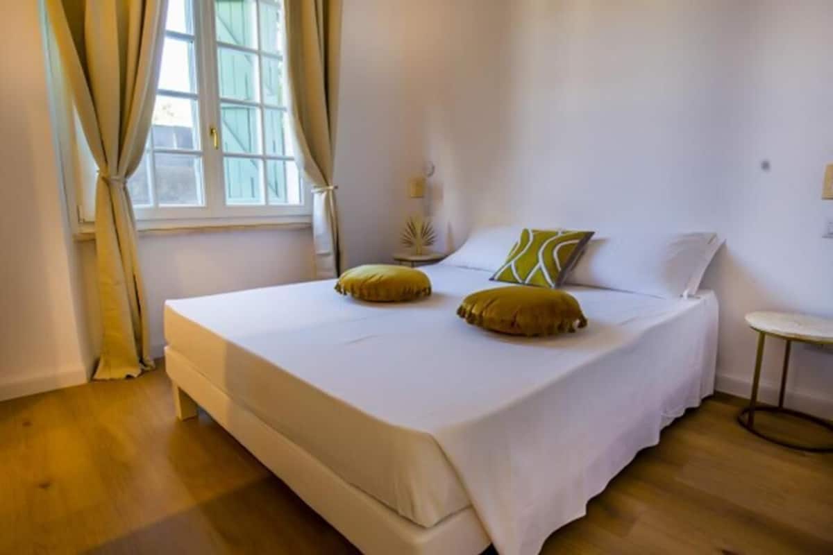 A bedroom with a bed, two pillows, a bedside table, and a window with curtains.