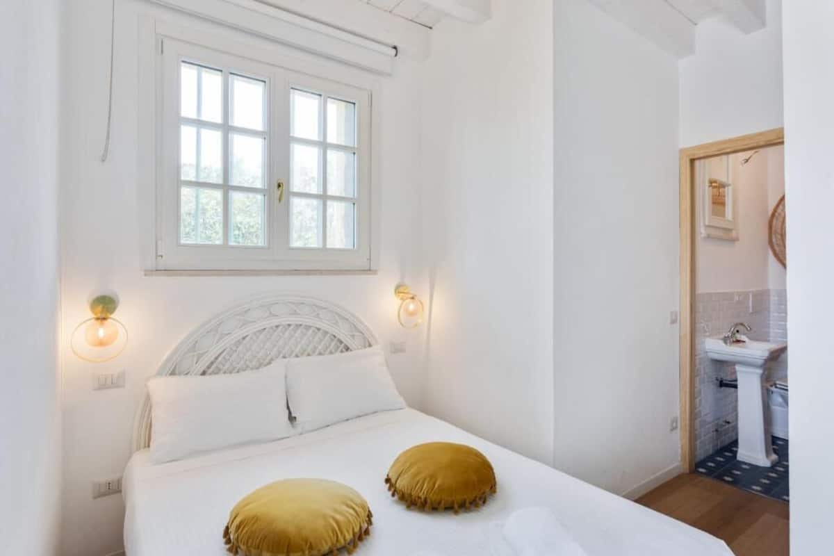 A bedroom with a bed, two bedside lamps, a window, and a view of a bathroom.