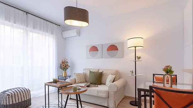 Una sala de estar moderna con un sofá, una mesa de centro y sillas. Hay una lámpara, un florero con flores y una obra de arte en la pared.