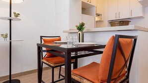 Un comedor con una mesa dispuesta para dos personas, sillas con cojines naranja, un jarrón con flores y una lámpara de pie.