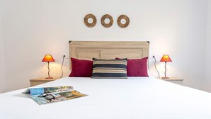 Una cama bien hecha con sábana blanca, dos almohadas rojas y una a rayas, dos lámparas de noche y tres adornos en la pared.