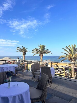 Een terras buiten met tafels en stoelen, palmbomen en uitzicht op zee.