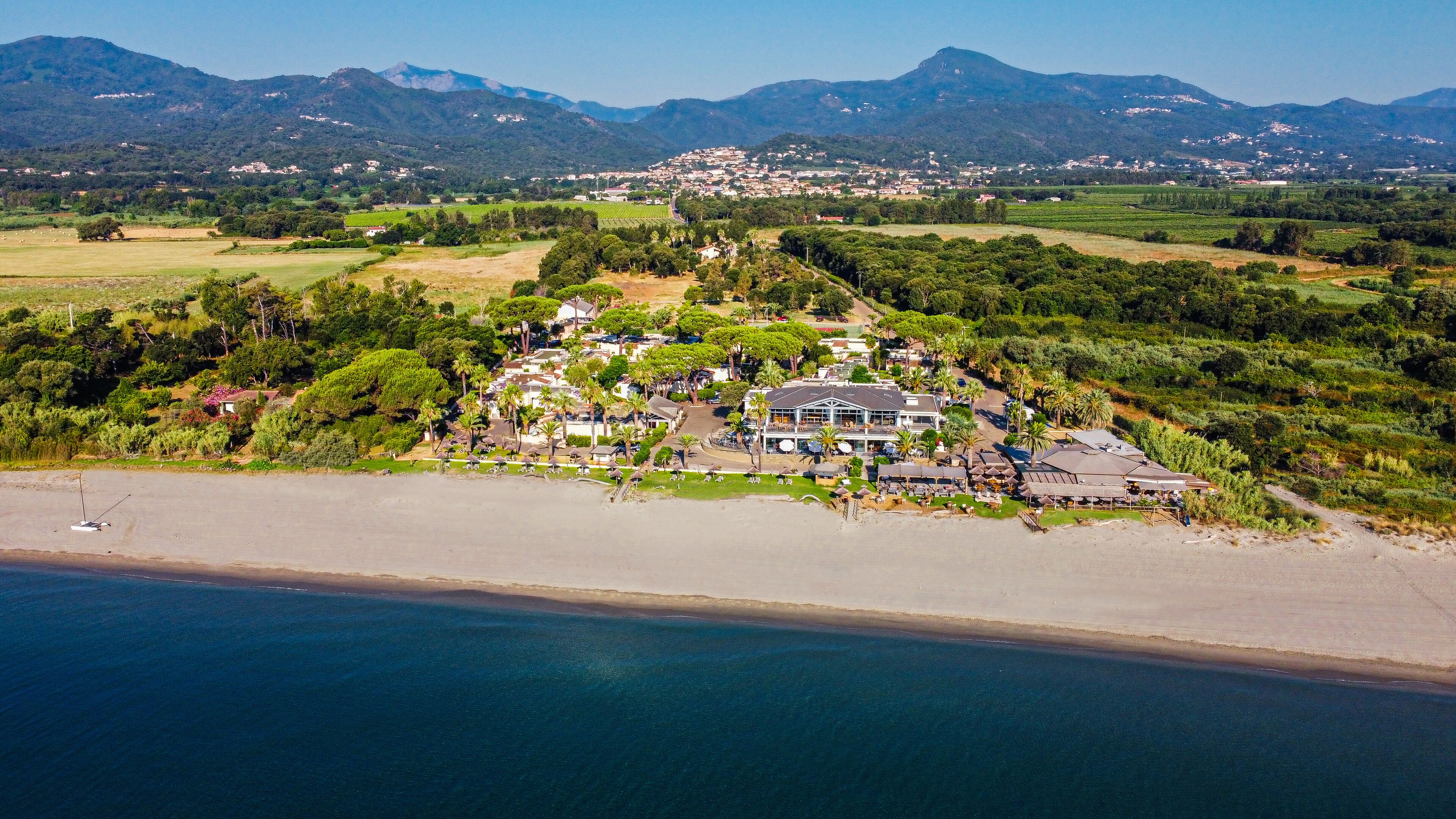 An aerial view of a coastal resort with a central building surrounded by lush greenery, a beach in the foreground, and mountains in the background.