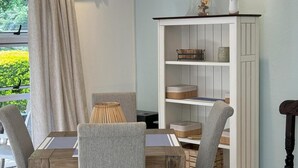 A dining area with a wooden table and grey upholstered chairs, a white bookshelf with decorative items, and a view of greenery outside. - Tranquil Cottage Retreat - Malanshof, Randburg (Johannesburg)