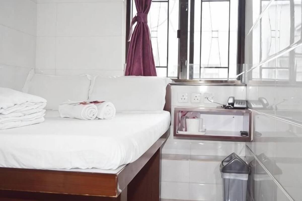 A hotel room with a bed, a nightstand with a phone and a lamp, and a window with a curtain.