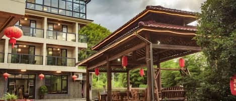 A traditional-style building with a covered outdoor seating area, surrounded by greenery and red lanterns.