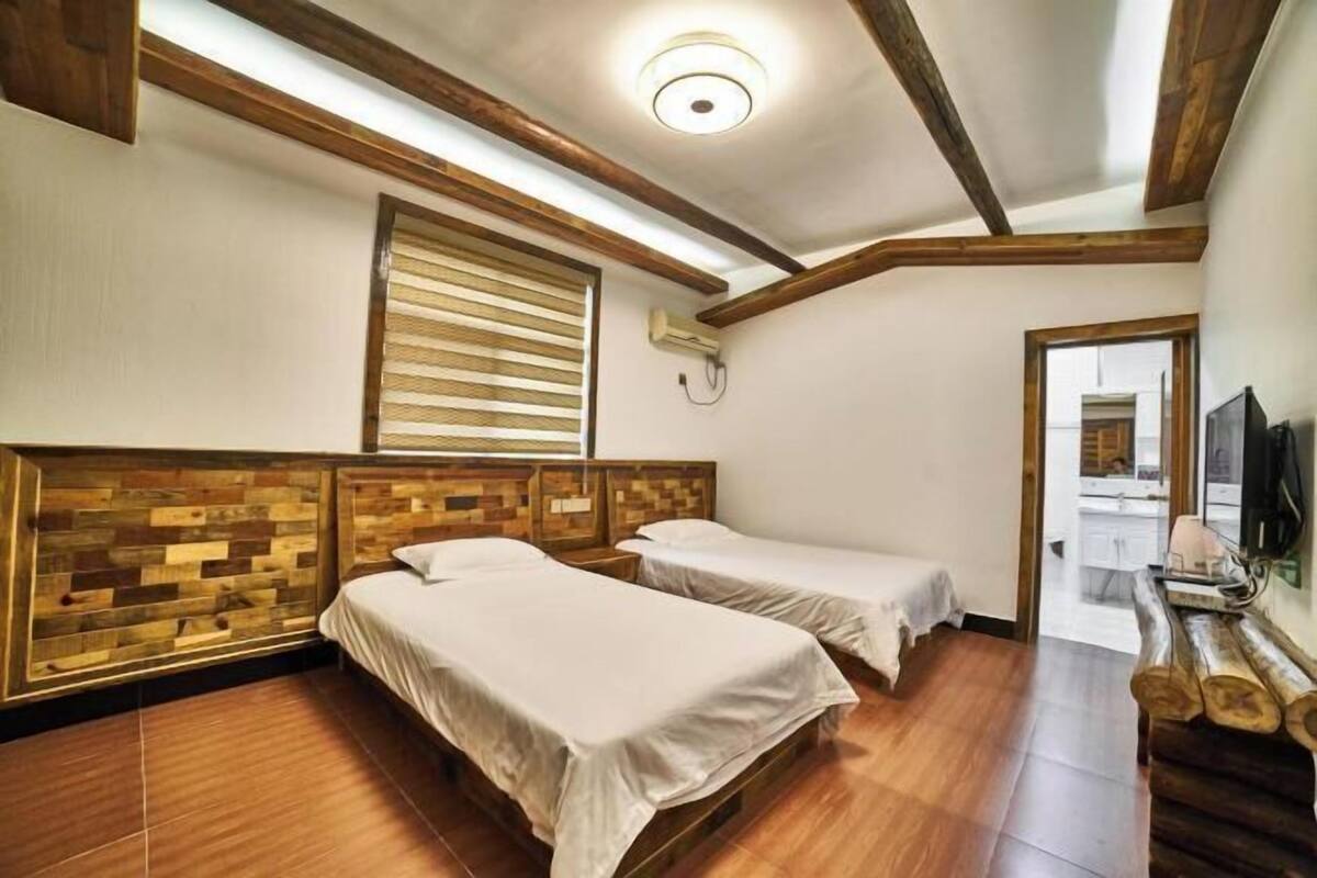 Une chambre avec deux lits, des têtes de lit en bois, un ventilateur de plafond, une fenêtre avec des stores et un téléviseur fixé au mur.