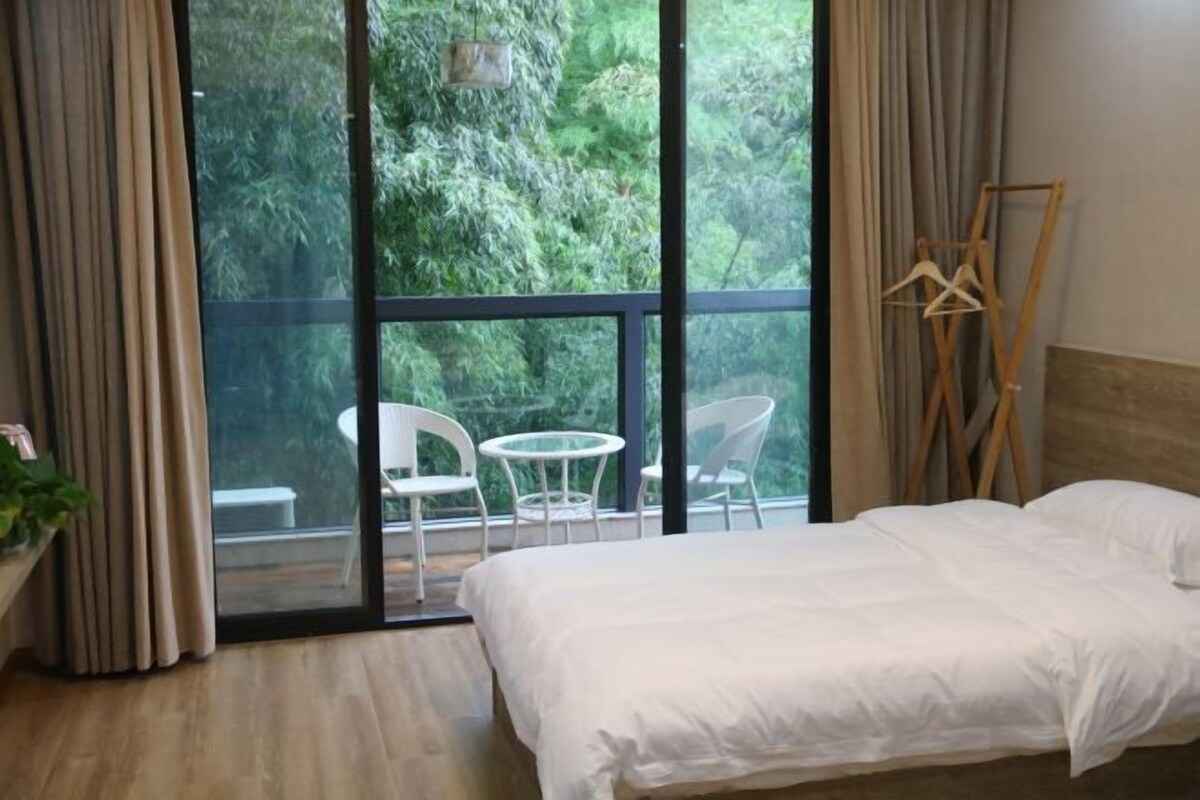 Une chambre avec un lit, une fenêtre donnant sur un paysage verdoyant, une petite table avec des chaises et un porte-manteau en bois.