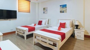 A hotel room with two single beds, a TV mounted on the wall, a small bedside table with a lamp, and a window with blinds. - RedDoorz Hostel @ Ritz Place Baguio (Baguio)