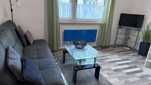 Ett modernt vardagsrum med en grå soffa, ett soffbord i glas, en blå bänk, en tv på ett tv-bord och en krukväxt.