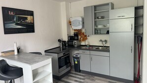 Ett kompakt kök med en gasspis, mikrovågsugn och ett kylskåp. Det finns en barstol och en papperskorg.
