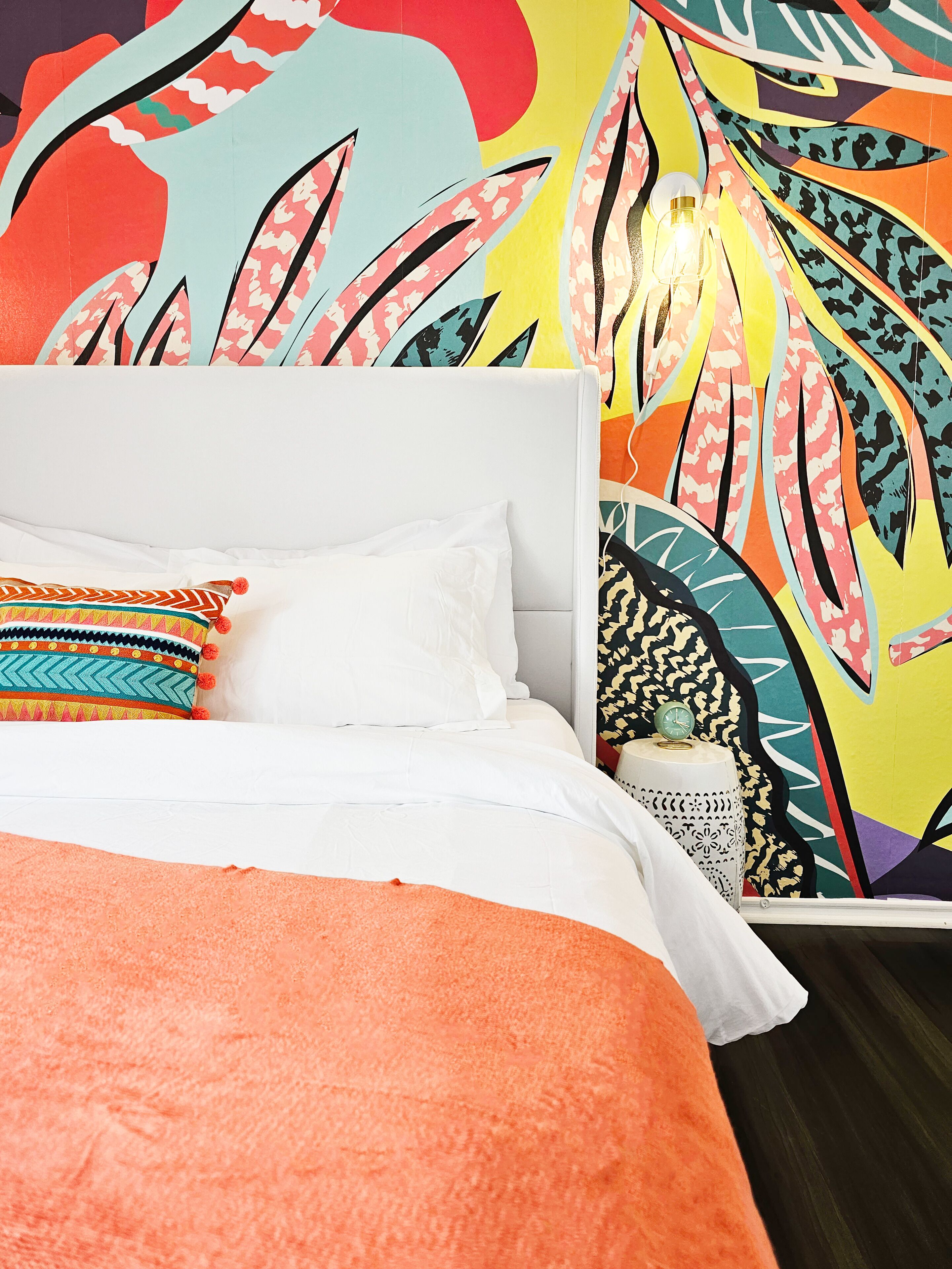 A bed with white bedding and colorful pillows, a patterned wall behind it, and a small bedside table with a clock and a decorative item.