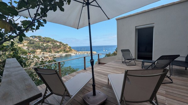 A wooden deck with outdoor furniture, including two lounge chairs and a table, overlooking a coastal area with boats and houses.