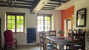 A dining area with a wooden table, chairs, and a red chair. There are windows with natural light, a chandelier, and a fireplace. - Gite "Le Chatel (Prissac)
