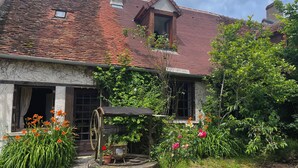 A rustic house with a traditional tiled roof, a wooden door, and a small garden with flowers and a water pump. - Gite "Le Chatel (Prissac)