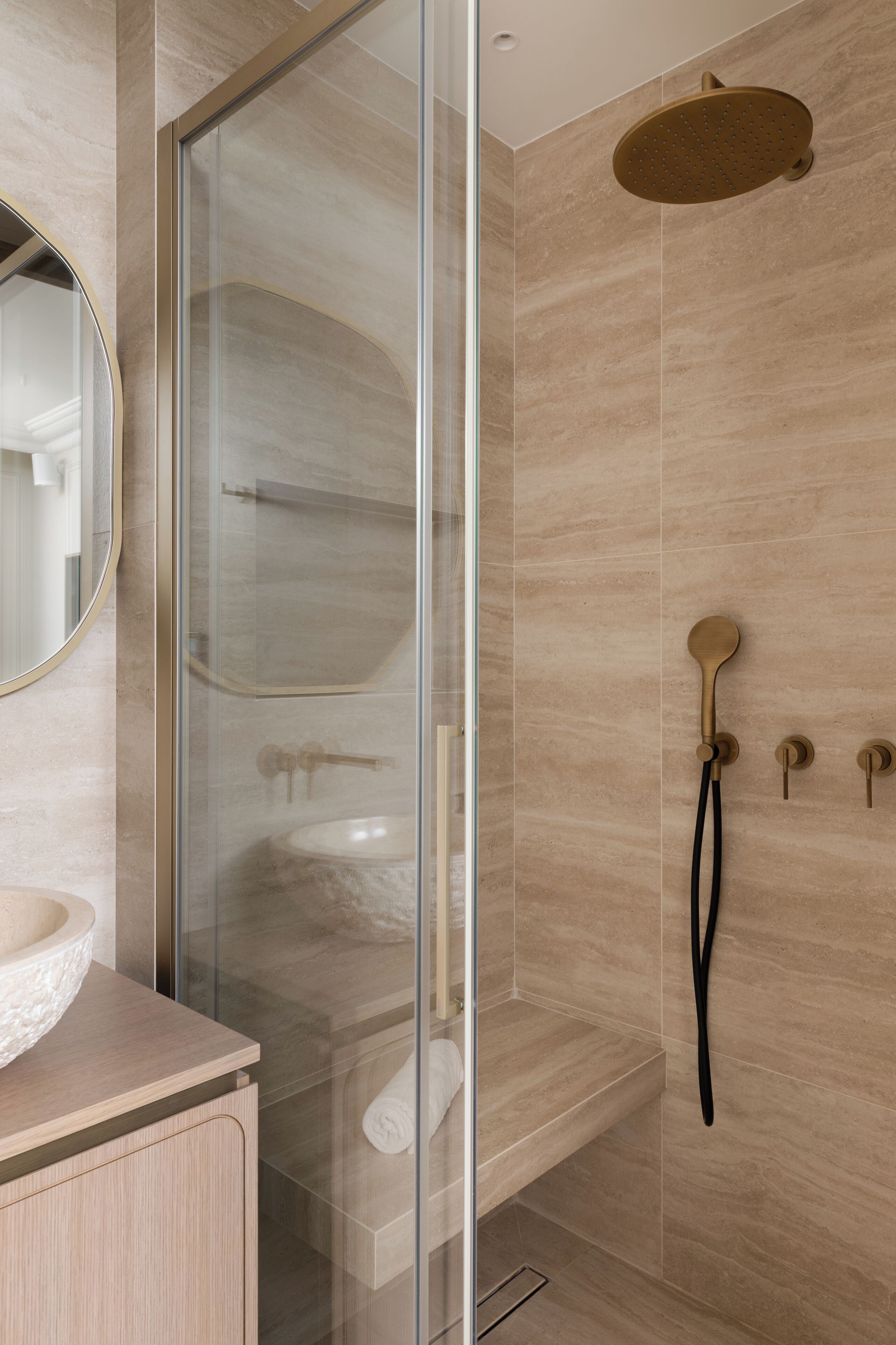 Un baño moderno con ducha envuelta en vidrio, un espejo redondo y pared revestida de azulejos color beige.