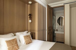 Una habitación de hotel con una cama grande, cabecera de madera, una lámpara de pared y un baño visible a través de una puerta abierta.