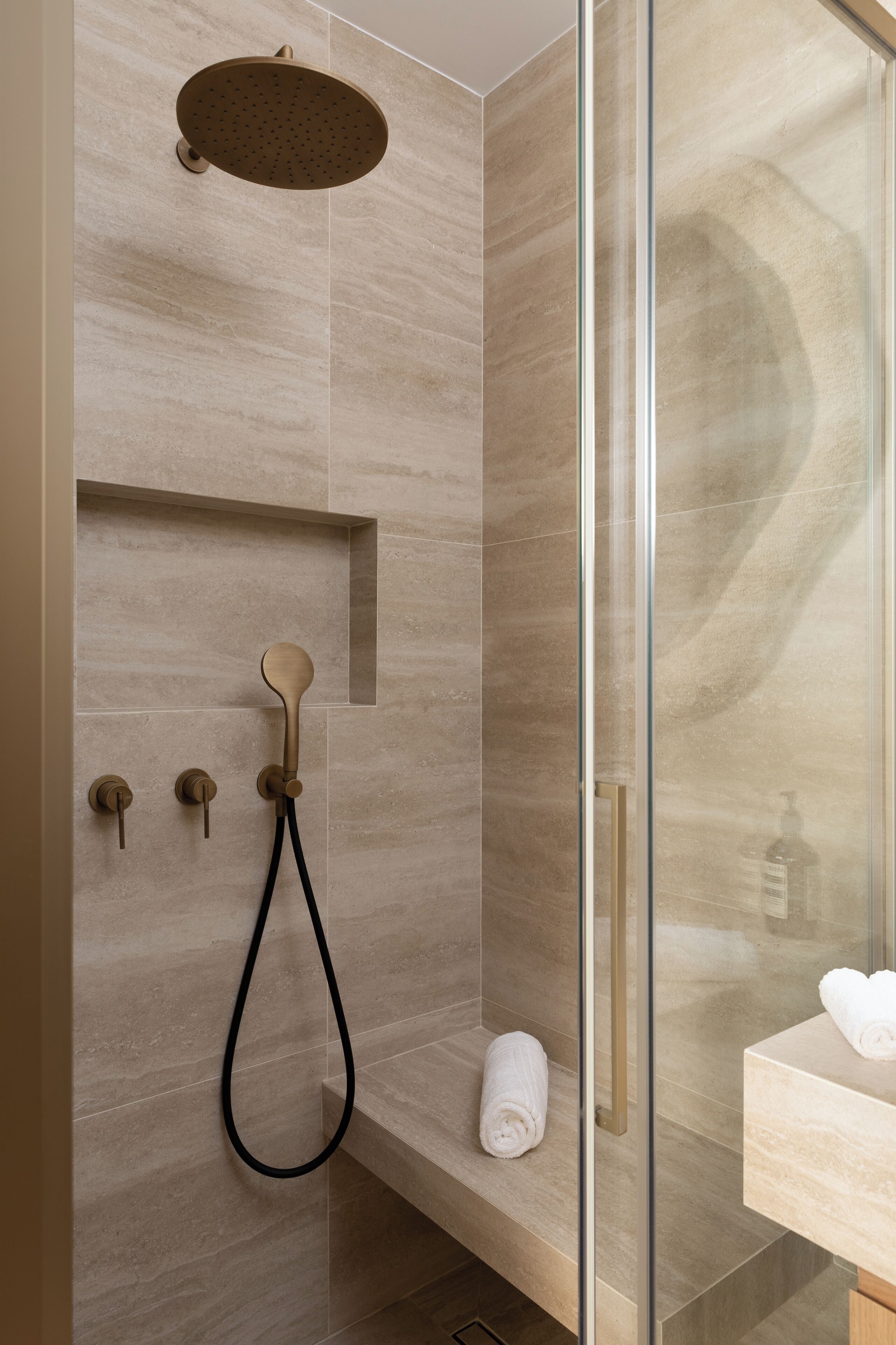 Una ducha moderna con cabezal de ducha manual y tipo lluvia integrado, cabina de ducha con mampara de vidrio y pared con azulejos de mármol.