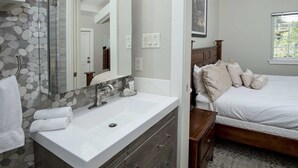Une salle de bain avec un lavabo blanc, un miroir et un mur en pierre. On y trouve un lit avec une tête de lit en bois et du linge de lit blanc.