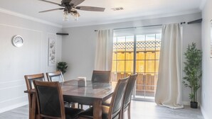 Un coin repas avec une table et des chaises en bois, un ventilateur de plafond, une horloge et une vue sur une clôture en bois, visible à travers une porte vitrée.