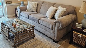 A living room with a grey sofa, a glass coffee table, and a rug. There are framed pictures on the wall and a sign that reads "BEACH HOUSE".