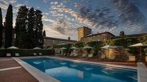 Piscina con sillas de descanso y sombrillas, rodeada de edificios de piedra y árboles.