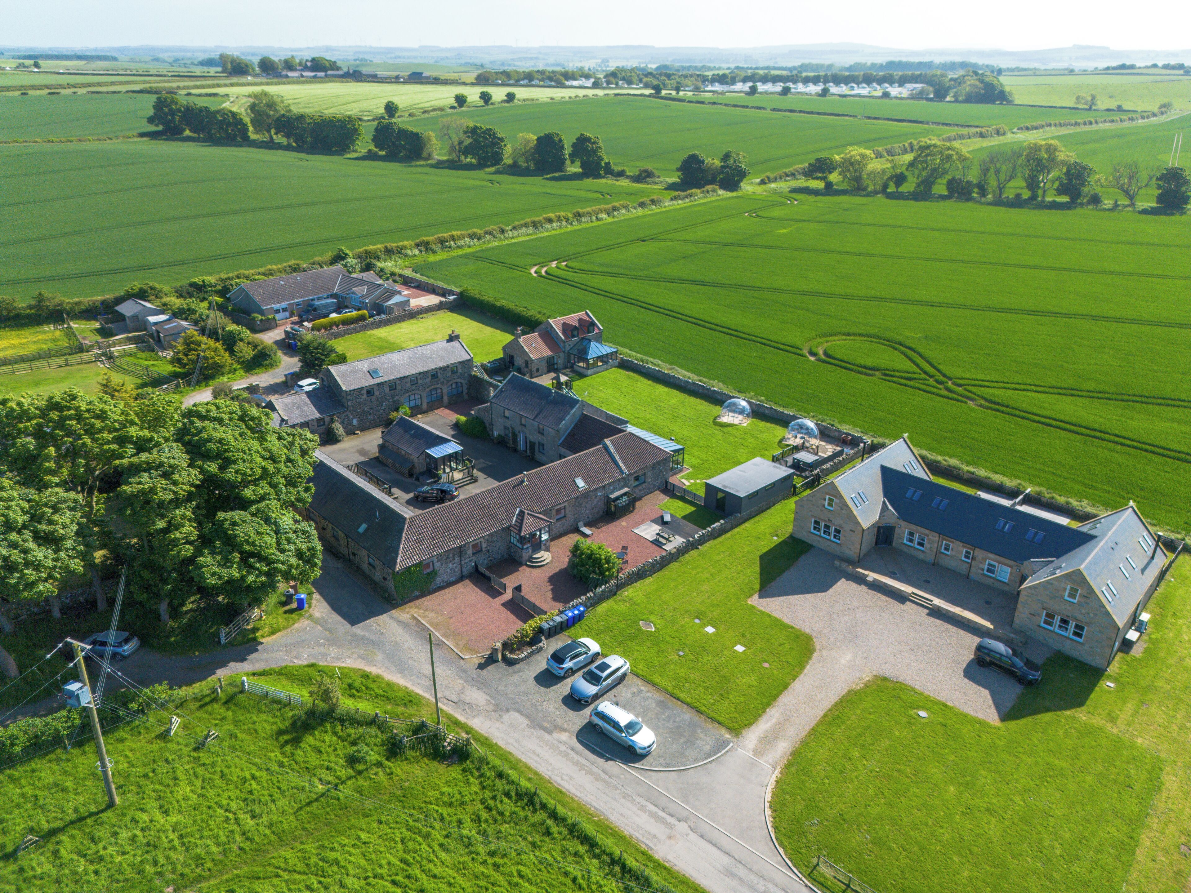 Vue aérienne d’une propriété rurale comprenant plusieurs bâtiments, un espace de stationnement et de vastes champs verdoyants.