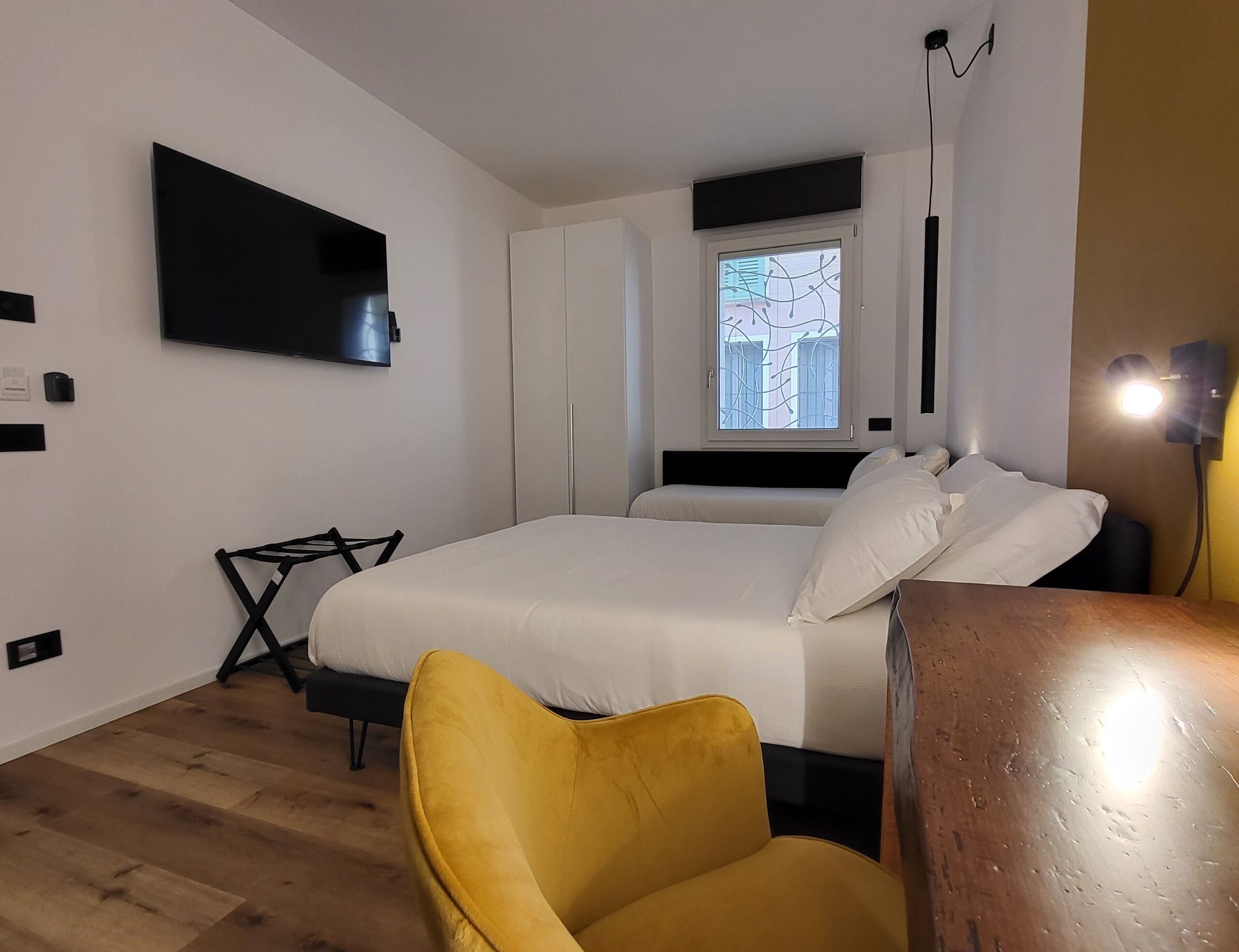 A modern bedroom with a bed, a yellow chair, a TV mounted on the wall, and a window with a view of buildings.