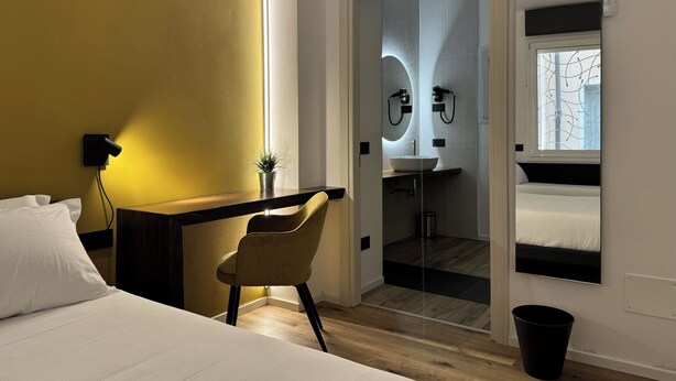 Une chambre d’hôtel moderne équipée d’un lit, d’un bureau, d’une chaise, d’une petite plante, d’une salle de bain avec un lavabo et un miroir, et d’une fenêtre.
