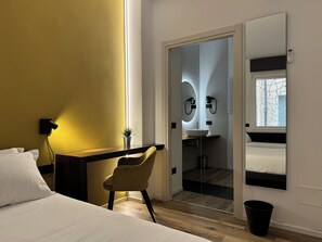 Une chambre d’hôtel moderne équipée d’un lit, d’un bureau, d’une chaise, d’une petite plante, d’une salle de bain avec un lavabo et un miroir, et d’une fenêtre.