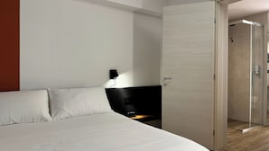 A modern hotel room with a large bed, a bedside table, a wall-mounted light, and a glass door leading to a shower area. - Delux Double Room in the center of Orzinuovi (Orzinuovi)