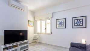 Ett vardagsrum med en platt-TV på ett vitt TV-bord, en vit stol, en grå soffa och två inramade lapptäcken på väggen.