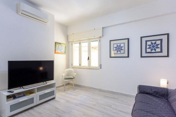 Una sala de estar con un televisor de pantalla plana sobre un pedestal blanco, una silla blanca, un sofá gris y dos colchas enmarcadas en la pared.
