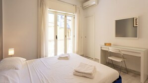 Ett hotellrum med en säng, ett skrivbord, en stol, en luftkonditionering och ett fönster med gardiner.