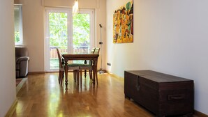 Ein Zimmer mit Holzboden, einem Esstisch mit Stühlen, einem dunklen Holzschrank und einem großen Fenster mit Jalousien.