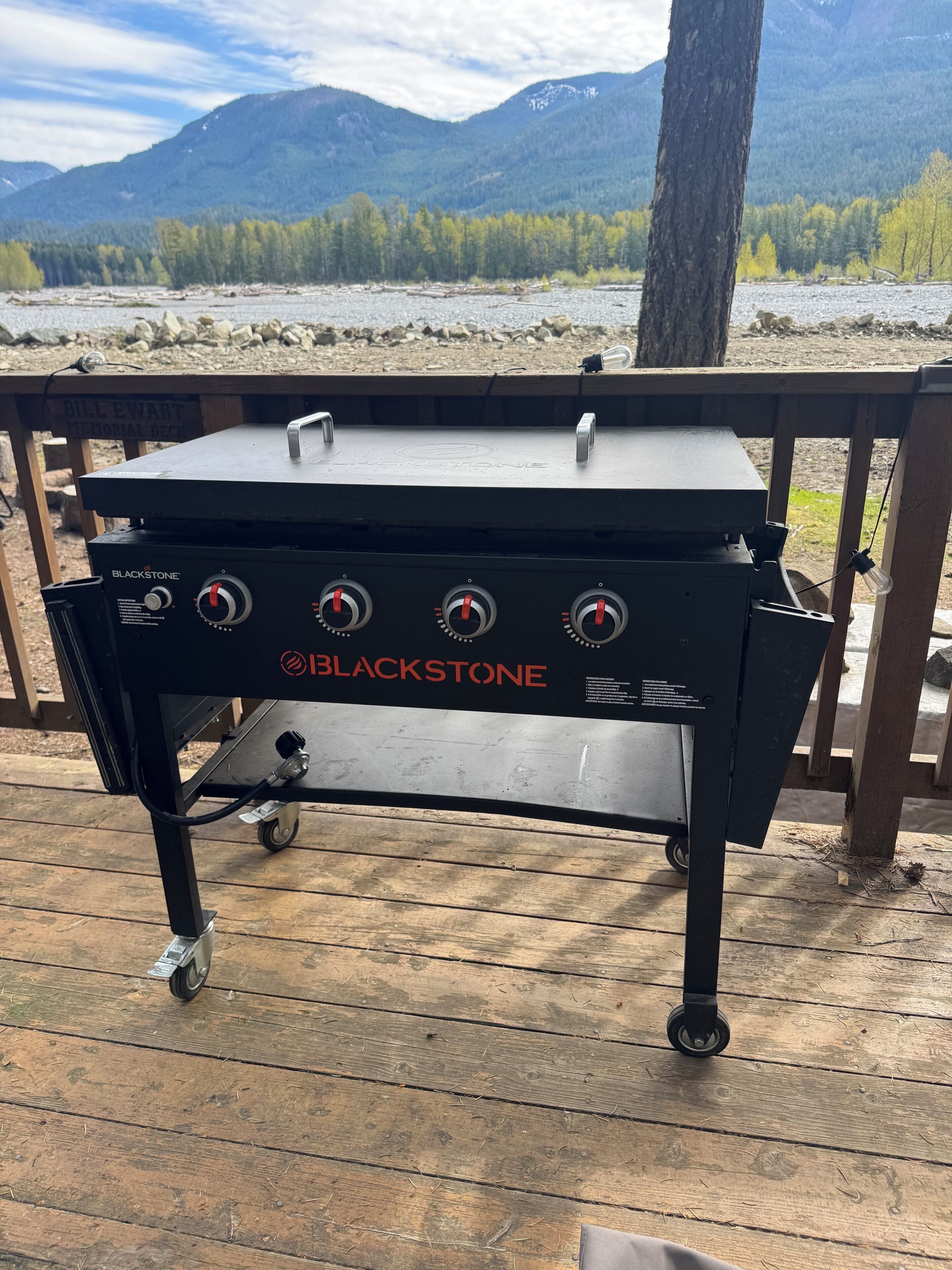 Una griglia Blackstone portatile su un pontile di legno, con vista sulle montagne e su un fiume.