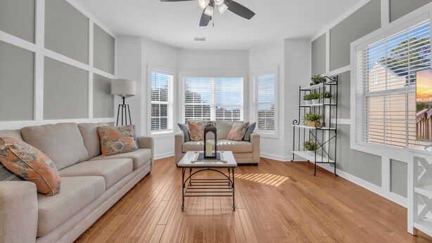 A living room with a beige sofa, a coffee table, and a ceiling fan. There are windows with blinds, a floor lamp, and a plant shelf. - Beach & Golf Vacation 2 (Myrtle Beach)
