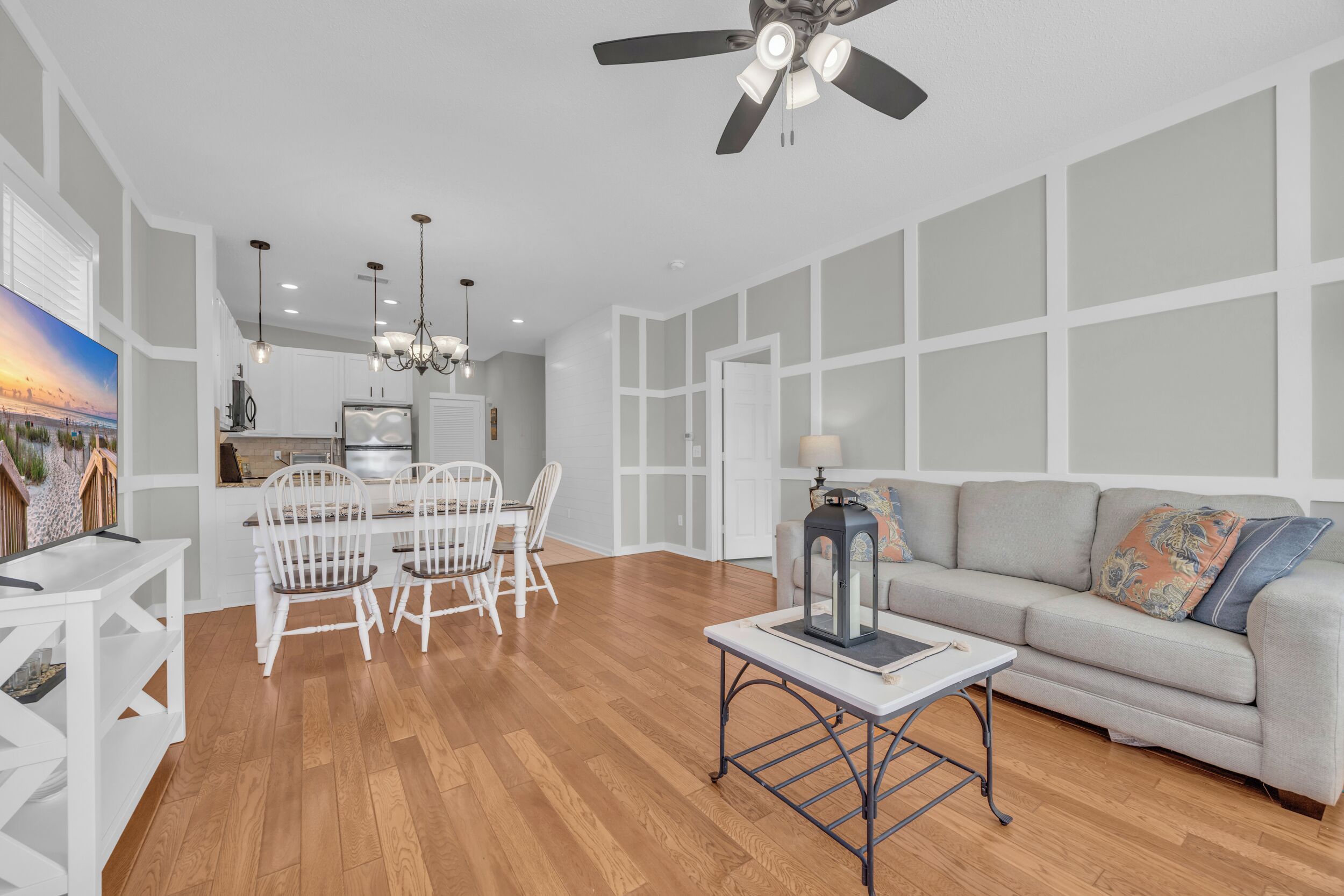 A modern living room with a grey sofa, white dining chairs, and a wooden floor. There is a ceiling fan, pendant lights, and a table with a lantern.