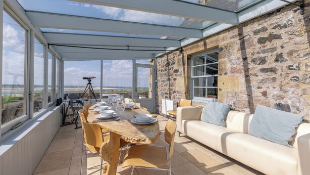 An outdoor dining area with a stone wall, a wooden table, and a white sofa. - Budle Newton - North East Escapes (Bamburgh)
