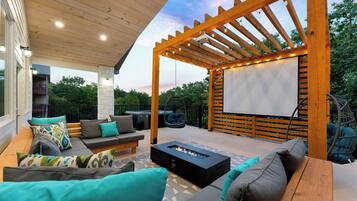 Une terrasse extérieure dotée d’une pergola en bois, d’un espace salon, d’un foyer et d’un grand écran pour les projections de films en plein air.