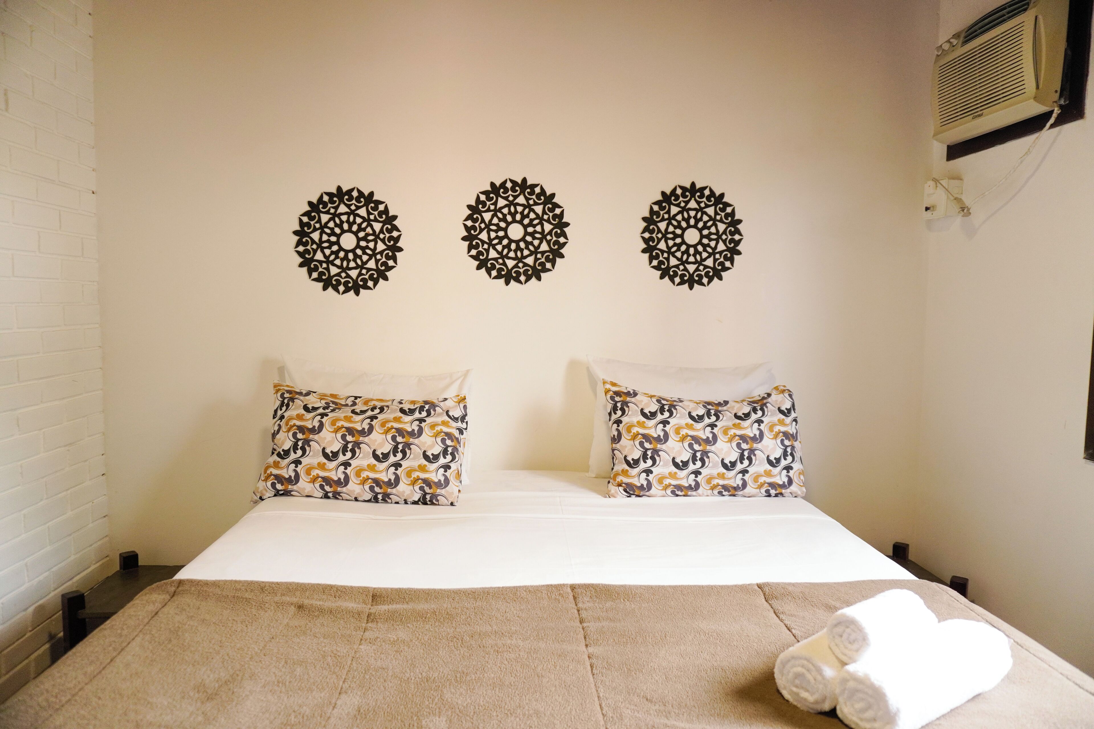 Habitación de hotel con cama, dos almohadones estampados, una colcha color beige y tres adornos en la pared.