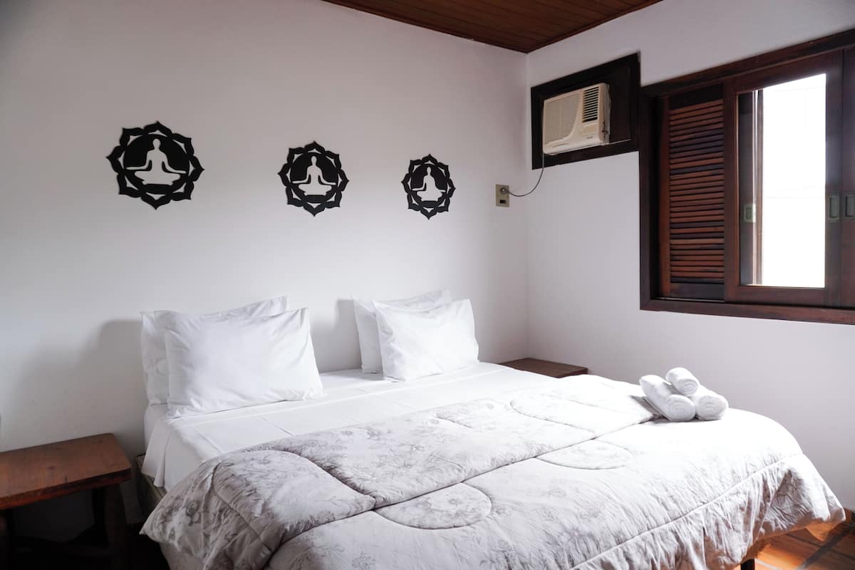 Ein ordentlich bezogenes Bett mit einem blumigen Bettbezug, weißen Kissen und einem Holzkopfende. Darüber hängen drei Wanddekorationen, und zur Rechten befindet sich ein Fenster mit Holzjalousien.
