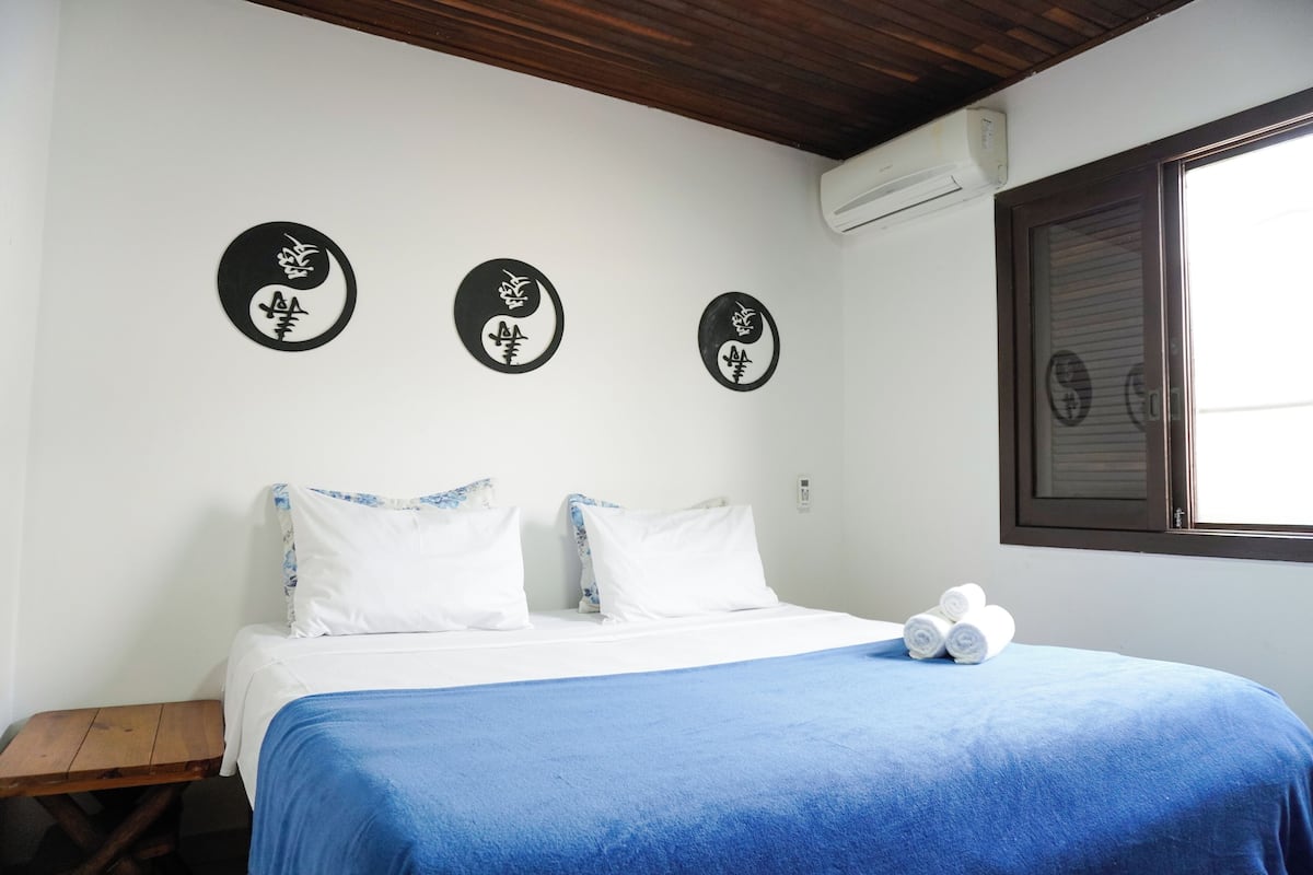 Ein ordentlich gemachtes Bett mit blauer Decke, weißen Kissen und einem Paar zusammengerollter Handtücher an der Seite. Drei schwarze, runde Wanddekorationen mit weißen Mustern sind zu sehen. Eine Holzbank ist links vom Bett sichtbar.