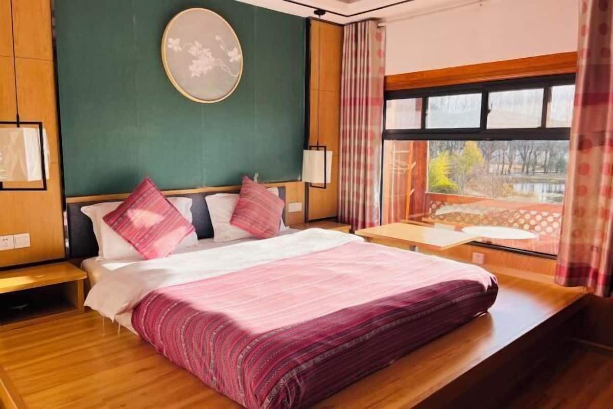 Une chambre d’hôtel comprenant un lit, une table de chevet, une fenêtre donnant sur des arbres et une œuvre d’art accrochée au mur.