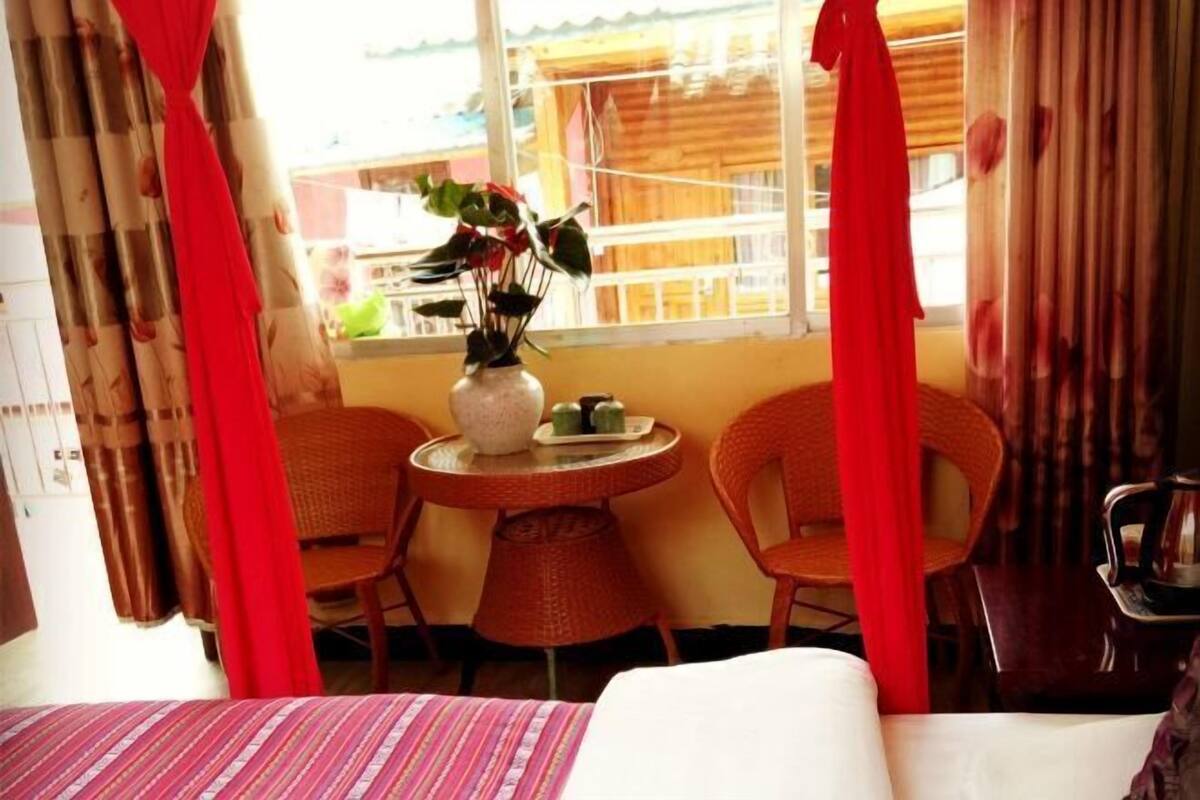 Une chambre d’hôtel avec un lit, une petite table sur laquelle se trouve un vase et deux tasses vertes, des chaises en osier et une vue par une fenêtre donnant sur un balcon en bois.