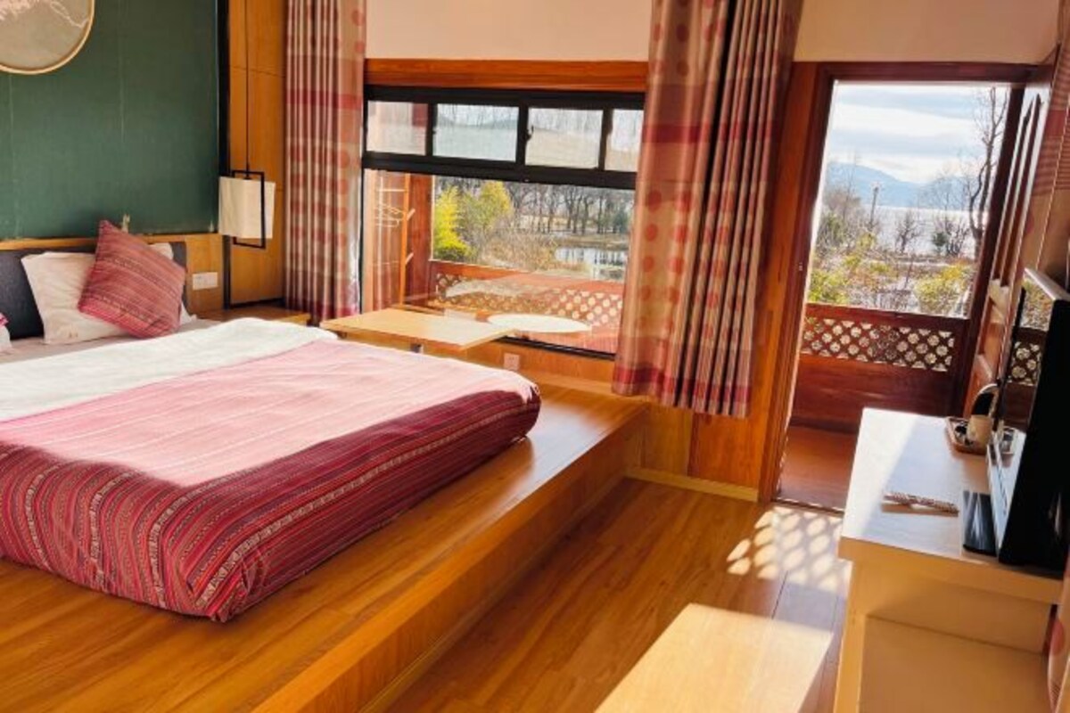 A bedroom with a bed, a desk, a window with curtains, and a view of the outdoors.