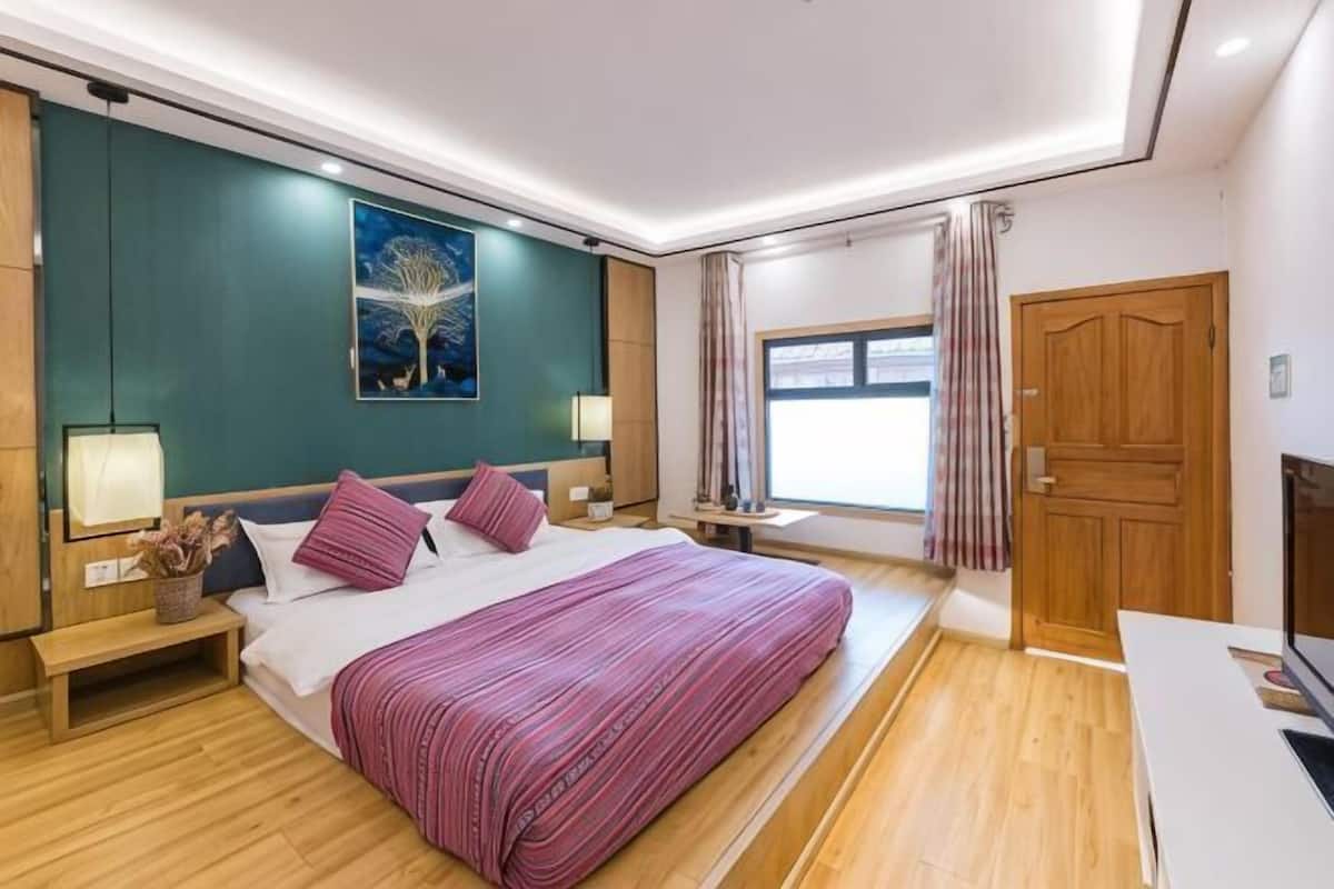 Une chambre d’hôtel moderne avec un grand lit, des tables de chevet, une petite table, une fenêtre avec des rideaux et une porte en bois.