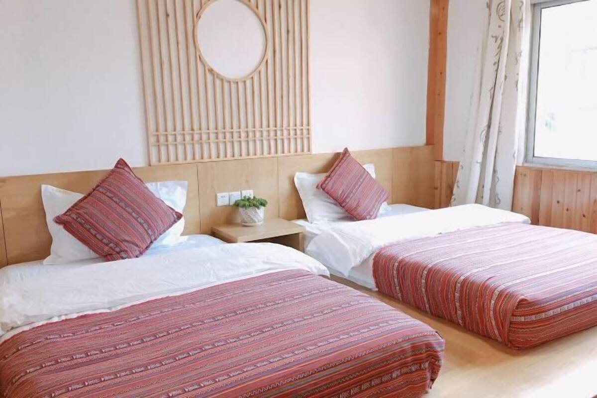 Une chambre double avec deux lits, chacun recouvert d’une couvre-lit à rayures rouges et agrémenté de coussins blancs. On y trouve une tête de lit en bois avec une découpe circulaire, une petite table de chevet avec une plante et une fenêtre avec des rideaux.