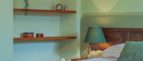 Una cama bien hecha con un almohadón verde, una lámpara de noche y una estantería con un reloj y objetos decorativos.