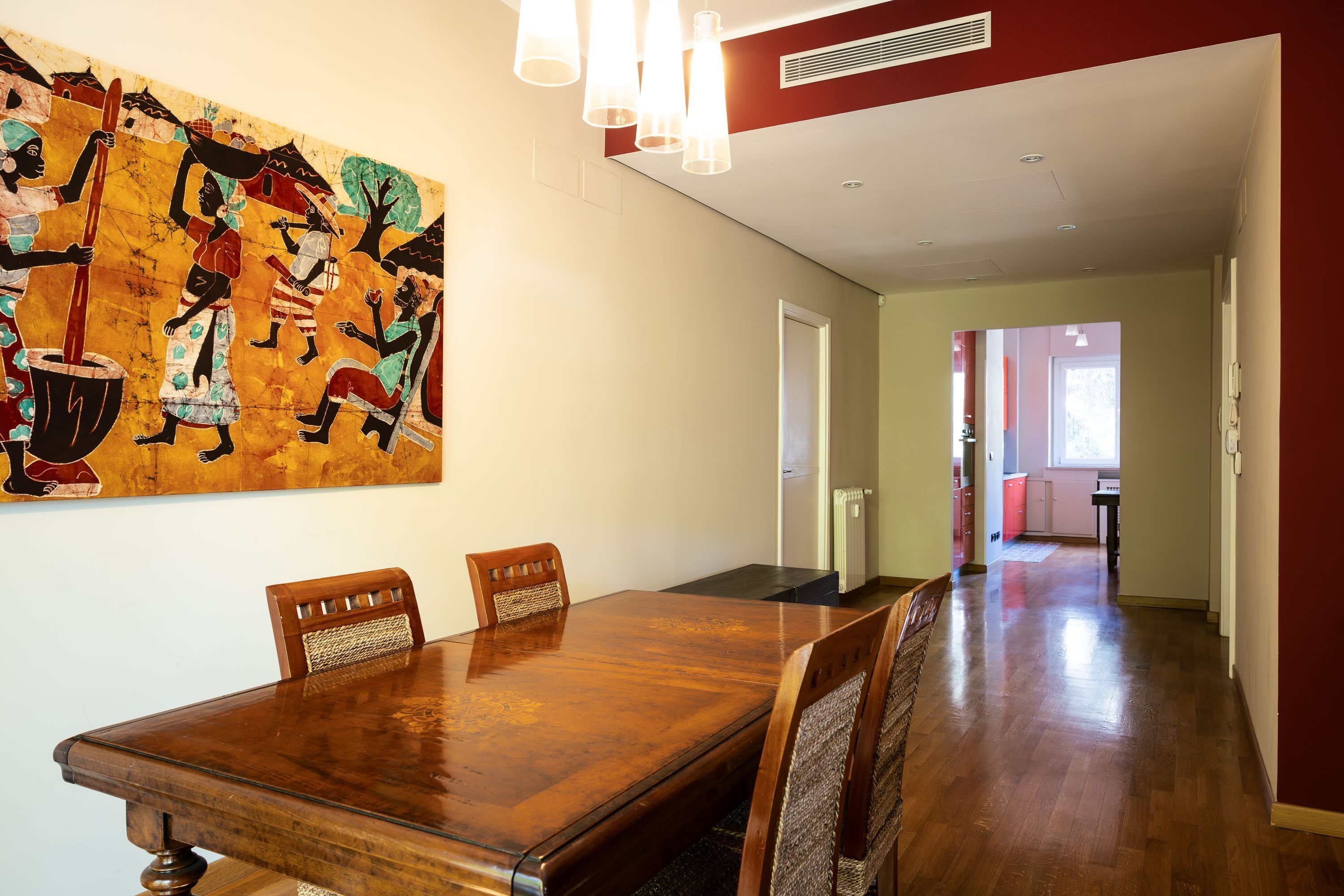 Un'area pranzo con tavolo e sedie in legno, un grande quadro decorativo a parete e un corridoio che conduce ad altre stanze.